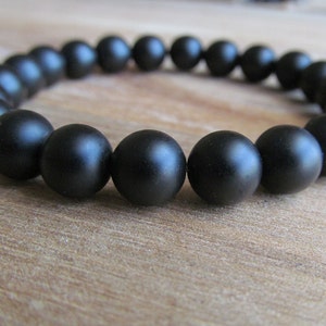 May include: A stretch bracelet composed of numerous matte black spherical beads arranged in a continuous circular loop. The beads have a smooth, non-reflective surface texture and are uniform in size, creating a consistent appearance around the entire circumference of the jewelry piece.
