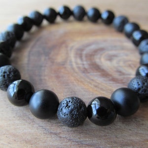 May include: A stretch bracelet composed of alternating black beads. The design features a mix of smooth, polished spherical beads and porous, textured volcanic rock beads arranged in a repeating pattern around the circular band.