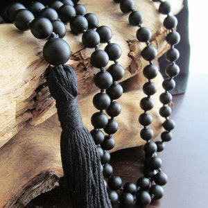 May include: A black beaded necklace with a tassel. The beads are matte and round. The necklace is draped over a piece of driftwood.