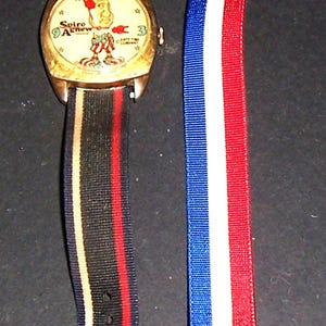 May include: Vintage wristwatch with a gold case and a cream face featuring a cartoon character and the text "Spiro Agnew". The watch has a black, red, and gold striped strap, and an additional red, white, and blue striped strap.