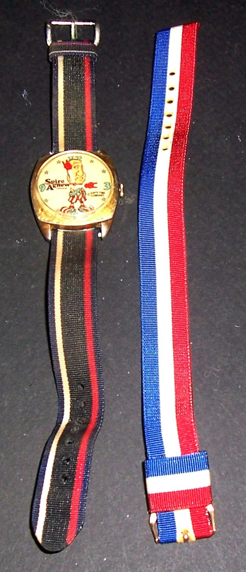 May include: Vintage wristwatch with a gold case and a cream face featuring a cartoon character and the text "Spiro Agnew". The watch has a black, red, and gold striped strap, and an additional red, white, and blue striped strap.