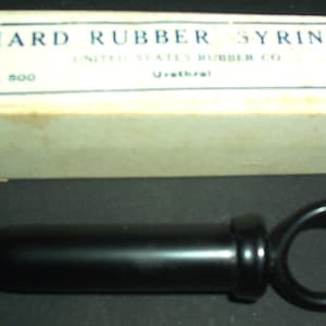 May include: A vintage hard rubber syringe with a black body and a ring handle, next to its original cardboard box. The box has blue text that reads "HARD RUBBER SYRINGE" and "UNITED STATES RUBBER CO."