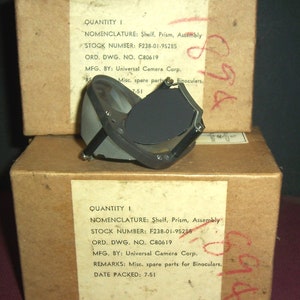 Puede incluir: Dos cajas de cartón con etiquetas que dicen "QUANTITY I" y "NOMENCLATURE: Shelf, Prism, Assembly". Las cajas contienen piezas de repuesto para binoculares. La caja superior tiene un ensamblaje de prisma de metal negro en la parte superior.