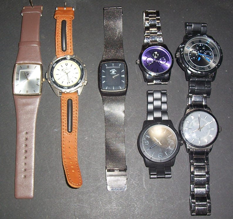 Pode incluir: Uma cole&ccedil;&atilde;o de sete rel&oacute;gios de pulso com designs variados, incluindo pulseiras de couro em castanho e bege, uma bracelete de malha preta e v&aacute;rias pulseiras de elos met&aacute;licos em tons de prata, preto e cinzento-a&ccedil;o com mostradores redondos e retangulares.