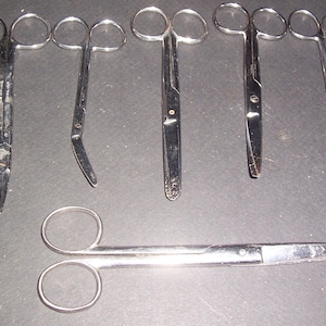 May include: A collection of six silver-colored metal surgical scissors arranged in a group. Five scissors are positioned vertically with finger loops at the top, while one pair of scissors is placed horizontally at the bottom of the frame.