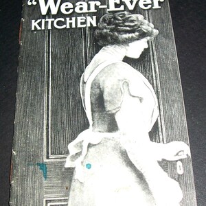 May include: Vintage book cover with the title "The Wear-Ever Kitchen" in white lettering. The cover features a black and white illustration of a woman in profile, standing near a door. The background is a dark, wood-grain texture.