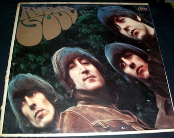 Puede incluir: Una funda de disco de vinilo cuadrada para el álbum Rubber Soul de The Beatles. La portada presenta una fotografía distorsionada en tonos cálidos de los cuatro miembros de la banda sobre un fondo oscuro y frondoso. El texto Rubber Soul está en letras marrones.