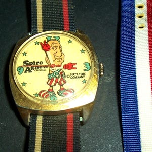 May include: Vintage gold wristwatch featuring a cartoon Spiro Agnew character on a cream-coloured face. The watch face has star accents and the text "Spiro Agnew Original" and "Dirty Time Company". The watch has a striped strap.