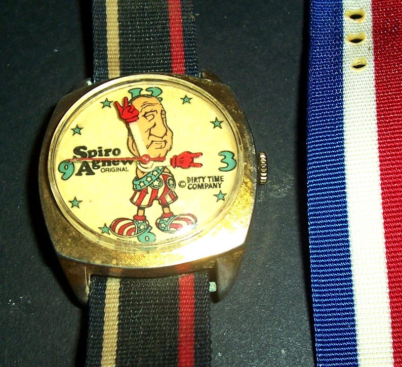 May include: Vintage gold wristwatch featuring a cartoon Spiro Agnew character on a cream-coloured face. The watch face has star accents and the text "Spiro Agnew Original" and "Dirty Time Company". The watch has a striped strap.