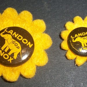 May include: Two round political campaign buttons featuring a dark brown background with yellow text reading LANDON KNOX and a yellow elephant graphic with GOP printed on it. Each button is mounted on a bright yellow felt flower-shaped backing.