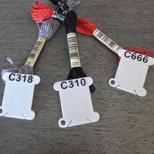 May include: Close-up of embroidery floss skeins in red, black, and gray. Each skein has a white label with a code: C318, C310, and C666. The floss is arranged on a gray surface, showcasing the vibrant colors and textures of the threads.