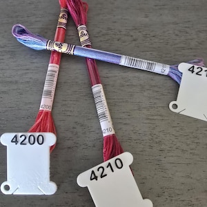 May include: Close-up of embroidery floss skeins in shades of red, purple, and blue. Each skein is labeled with a white tag displaying a number. The floss is arranged on a gray surface, showcasing the vibrant colors and textures of the thread.