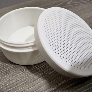 May include: A white, round container with a matching lid. The lid is perforated with numerous small holes. The container is open, revealing its interior. The items are made of a smooth, matte material.