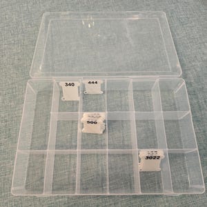 May include: A clear plastic organiser box with a hinged lid, divided into multiple compartments. The box contains small spools of thread labelled with numbers 340, 444, 500, and 3022. The box is open and sits on a light blue surface.
