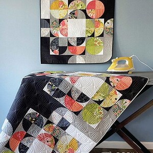 May include: A quilt with a "Partial Eclipse" design, featuring semi-circular patterns in black, white, gray, yellow, green, and red. The quilt is displayed on a table with a yellow iron. The text "Partial Eclipse" is at the top.