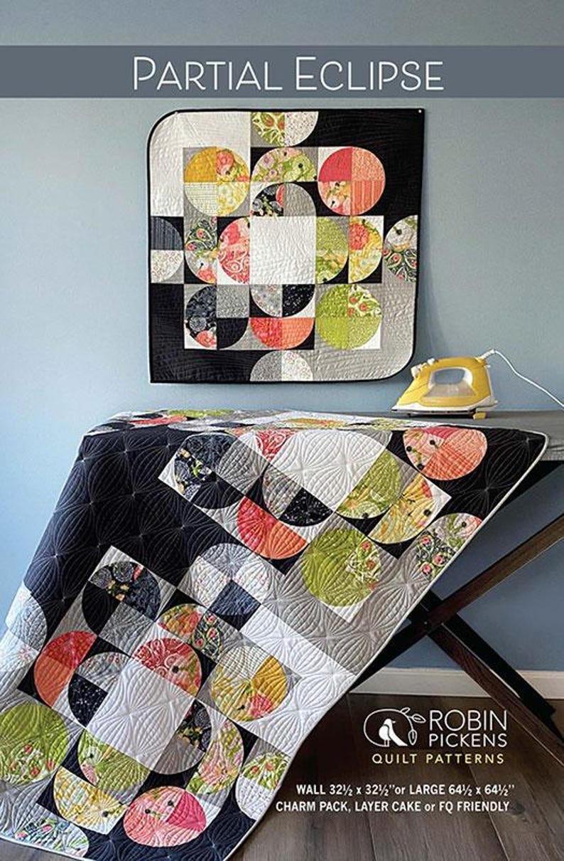 May include: A quilt with a "Partial Eclipse" design, featuring semi-circular patterns in black, white, gray, yellow, green, and red. The quilt is displayed on a table with a yellow iron. The text "Partial Eclipse" is at the top.