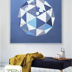 May include: A framed quilt with a geometric design in shades of blue and white hangs on a wall. The quilt's design resembles a globe. A navy blue bench with a mustard yellow throw is below the quilt. The word "GLOBE" is visible.