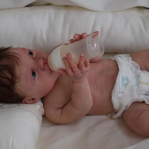 Puede incluir: Una muñeca realista de recién nacido con cabello castaño oscuro y ojos azules, acostada boca arriba. La muñeca lleva un pañal blanco y sostiene un biberón de plástico transparente con tapa blanca en la boca, simulando alimentarse sobre una almohada.