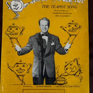 Könnte beinhalten: Ein Vintage-Notenblatt mit einem Cartoon-Teekanne mit einem lächelnden Gesicht und dem Titel "She's a Little Tea Pot, The Teapot Song". Das Cover zeigt auch ein Foto eines Mannes im Anzug und mit Fliege, sowie den Text "Words and Music by Clarence Kelley and Geo. H. Sanders".