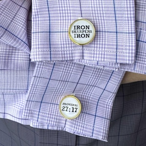 Puede incluir: Primer plano de una camisa a cuadros de color púrpura claro con gemelos dorados. Los gemelos tienen un fondo blanco con el texto "IRON SHARPENS IRON" y "PROVERBIOS 27:17" en negro. La camisa tiene un patrón de cuadrícula azul.
