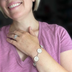 May include: A silver charm bracelet with multiple round charms featuring inspirational quotes. The charms are connected by a silver chain. The bracelet is worn on a person's wrist.