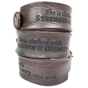 May include: Three stacked brown leather bracelets. Each bracelet is embossed with the words "She is clothed with Strength & Dignity" and "PROV. 31:25". The bracelets are fastened with a buckle.