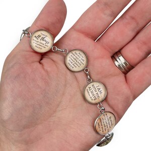 May include: A silver bracelet with five round glass charms. Each charm features a different Bible verse in a vintage style font on a beige background. The verses are: "I can do all things through Christ, who strengthens me. Phil 4:13", "And my God shall fulfill every need of yours according to his riches in glory in Christ Jesus. Phil 4:19", "I am with you always. Matthew 28:20", "Be strong and courageous. The Lord your God will be with you wherever you go. Joshua 1:9"