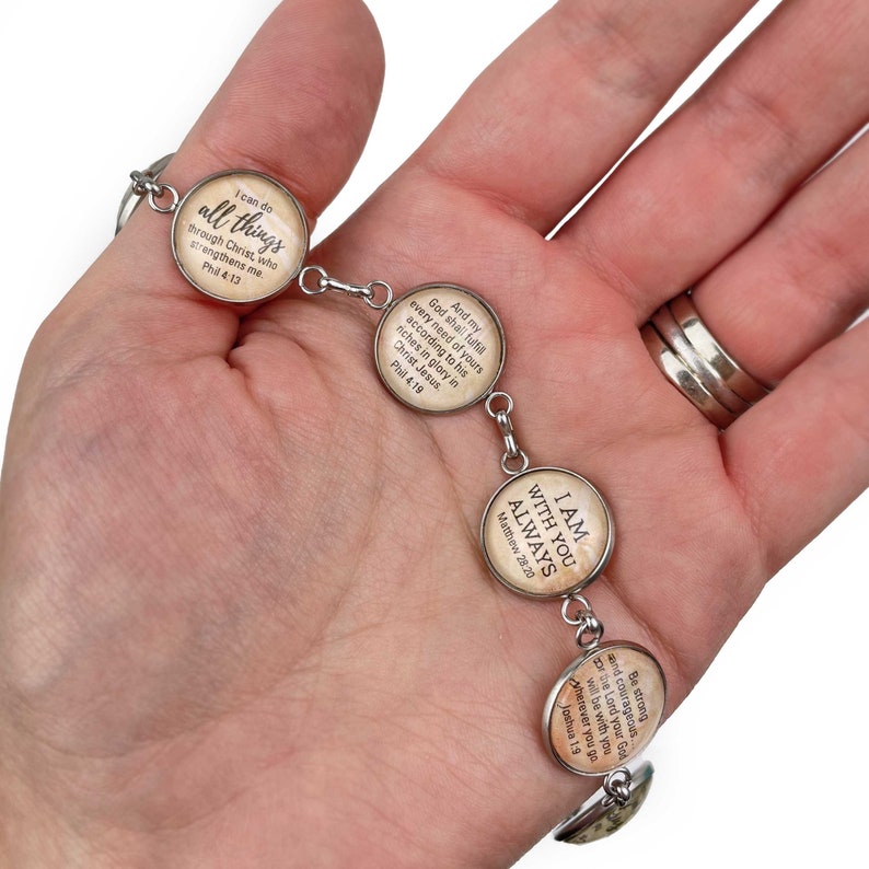 May include: A silver bracelet with five round glass charms. Each charm features a different Bible verse in a vintage style font on a beige background. The verses are: "I can do all things through Christ, who strengthens me. Phil 4:13", "And my God shall fulfill every need of yours according to his riches in glory in Christ Jesus. Phil 4:19", "I am with you always. Matthew 28:20", "Be strong and courageous. The Lord your God will be with you wherever you go. Joshua 1:9"