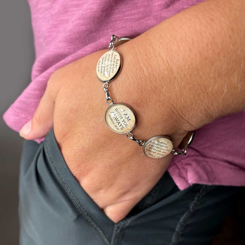May include: A silver bracelet with four oval charms. Each charm has a different bible verse printed on it. The bracelet is on a person's wrist.