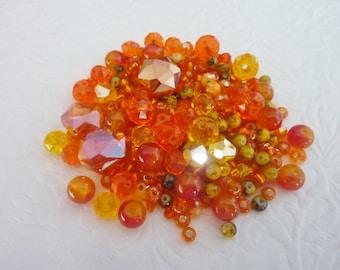 May include: A collection of orange and yellow beads in various shapes and sizes. The beads include faceted crystals, round beads, and smaller seed beads. The colors range from light yellow to deep orange and red, with some iridescent highlights.