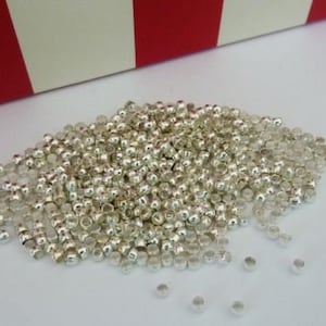 May include: A collection of small, shiny silver beads scattered on a white surface. The beads are round with a hole through the centre. A red and white striped box is in the background.