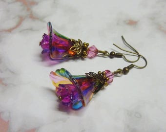 May include: A pair of handcrafted earrings featuring colorful, iridescent flower-shaped pendants. The earrings have a bronze-colored metal with pink bead accents and fish hook closures. The flowers display shades of purple, pink, yellow, and blue.