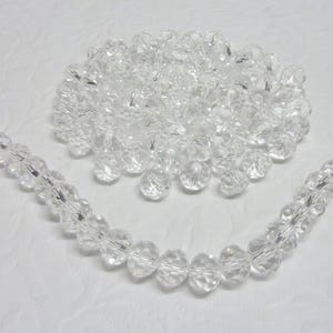 May include: A strand of clear crystal beads, with a pile of loose beads in the background. The beads are faceted and have a round shape.