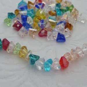 May include: A selection of multi-coloured crystal beads, faceted and in various shapes. Some are scattered, others strung together in a curved line. The colours include red, blue, green, and yellow, creating a rainbow effect.