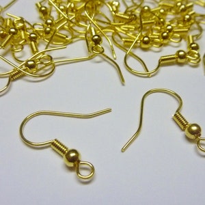 May include: A close-up of a pile of gold-toned earring hooks. The hooks are made of metal and have a small loop at the top. The hooks are scattered on a white surface.