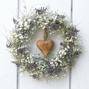 Könnte beinhalten: Ein kreisförmiger Kranz aus weißen und lila Blüten mit einem Holzherz-Anhänger. Der Kranz besteht aus getrockneten Blumen und Zweigen und hängt vor einem weißen Hintergrund. Das Herz ist hellbraun und wird von einem Naturfaden gehalten.