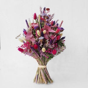 Könnte beinhalten: Ein lebendiger Strauß aus getrockneten Blumen in Rosa-, Lila- und Cremetönen. Das Arrangement weist verschiedene Texturen und Formen auf, die mit Bindfaden zusammengebunden sind. Die Blumen umfassen Lavendel, Disteln und andere Wildblumen.