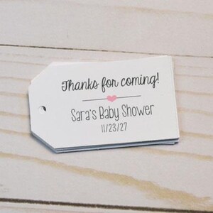 Könnte beinhalten: Ein Stapel weißer Geschenkanhänger mit dem Text "Thanks for coming!" über "Sara's Baby Shower" und dem Datum "11/23/27". Ein kleines rosa Herz befindet sich zwischen den Sätzen. Die Anhänger haben ein kleines Loch auf der linken Seite.