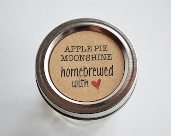 Könnte beinhalten: Ein silberner Einmachglasdeckel mit einem braunen Etikett, auf dem "APPLE PIE MOONSHINE homebrewed with" und ein rotes Herz steht.