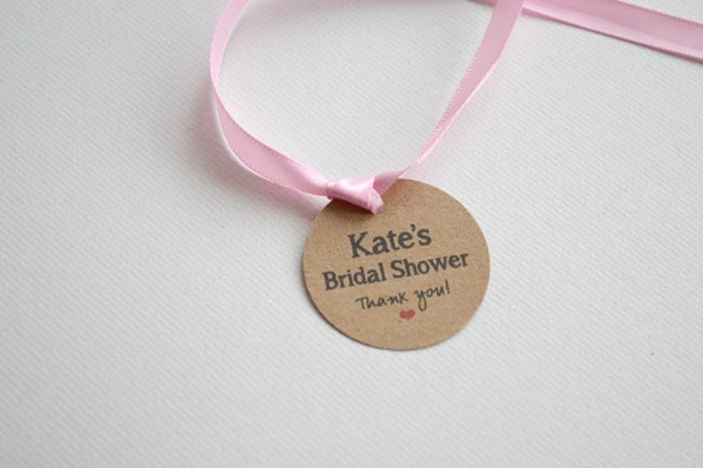 May include: A brown circle tag with a pink ribbon attached. The tag says "Kate's Bridal Shower Thank you!" with a small red heart.