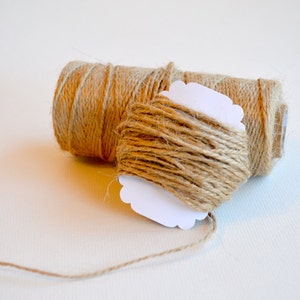 May include: Two spools of natural brown twine with a white paper tag attached to one spool. The twine is unwinding from the spool.