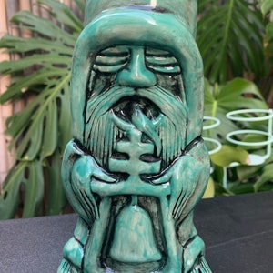 May include: A teal green ceramic tiki mug shaped like a person with a long beard and a closed-eye expression. The person is holding a large, round object in their hands.