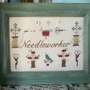 May include: A framed needlepoint artwork with a green wooden frame. The artwork features the word "Needleworker" in a script font, along with cross-stitched images of flowers, spools of thread, scissors, and a bird. The color palette includes beige, brown, and blue.