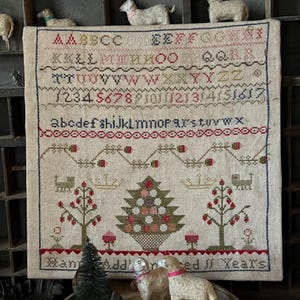 Può includere: Un ricamo a punto croce beige con un disegno di albero di Natale. Il ricamo include l'alfabeto, i numeri e il testo "We've Added 11 Years".