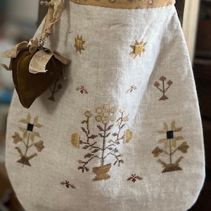 May include: A white linen bag with a brown trim and embroidered floral designs in gold and brown. The bag has a small brown velvet heart attached to it.