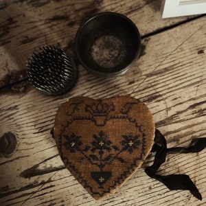 May include: A brown heart-shaped pincushion with black embroidery of flowers and a crown. The pincushion has a black ribbon attached to it.