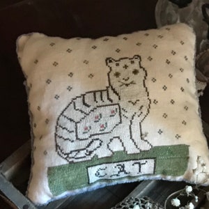 Può includere: Un cuscino bianco con uno sfondo verde e un gatto bianco con una striscia marrone sulla schiena. Il gatto è seduto su una base verde con la parola "GATTO" ricamata su di essa. Il cuscino ha uno sfondo bianco con un piccolo motivo a stella bianca.