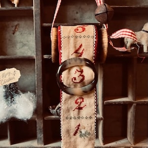 May include: A vintage-style Christmas countdown ribbon with red and white gingham ribbon, a wooden spool, and a brown ring with the numbers 1 through 5 embroidered on it. The ribbon is hanging from a wooden drawer unit.