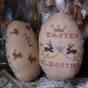 May include: Two beige, egg-shaped ornaments with cross-stitched designs. One features birds, flowers, and bunnies. The other says "EASTER BLESSING" with a crown and bunnies. The ornaments are likely for Easter.