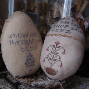 Può includere: Due uova di stoffa bianca con disegni ricamati. Un uovo ha il testo "Mostrami la via giusta O Signore" e l'altro ha un disegno floreale con fiori rossi.